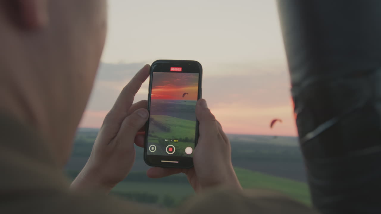 Rear view of lady recording scenic sunset and distant paraglider using smartphone while riding in hot air balloon over countryside, glowing sky and open landscape