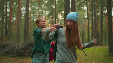 young woman in green helps her friend put on a bag as they stand in a serene forest setting, with warm sunlight casting a glow in the background