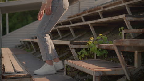 close-up of girl s legs as she steps up rustic stadium seating, she sat on the first roll, with visible greenery growing among rusty seat frames, background shows blurred greenery