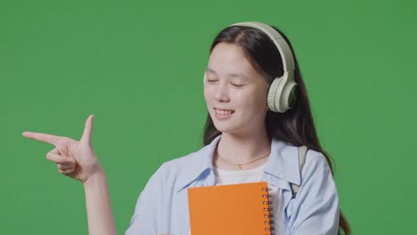 close up of asian teen girl student with a backpack and some books smiling and pointing to side while standing in the green screen background studio