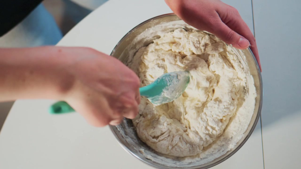 Hands mix dough in metal bowl with green spatula. Person focuses on blending ingredients smoothly. Scene shows baking preparation in clean kitchen setting. Ideal for cooking, recipe