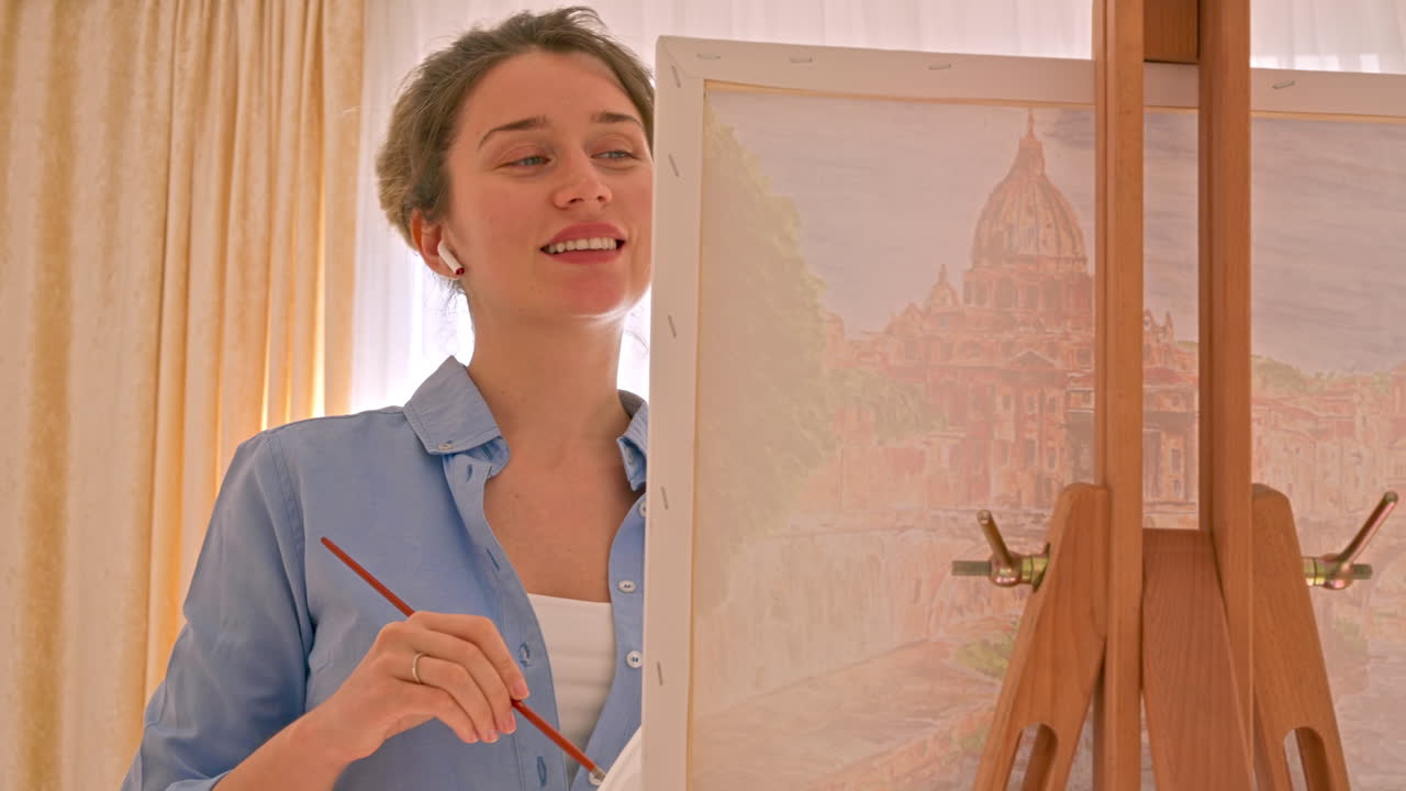 Caucasian woman painting a picture with a palette in her hands and listening music with earphones indoors