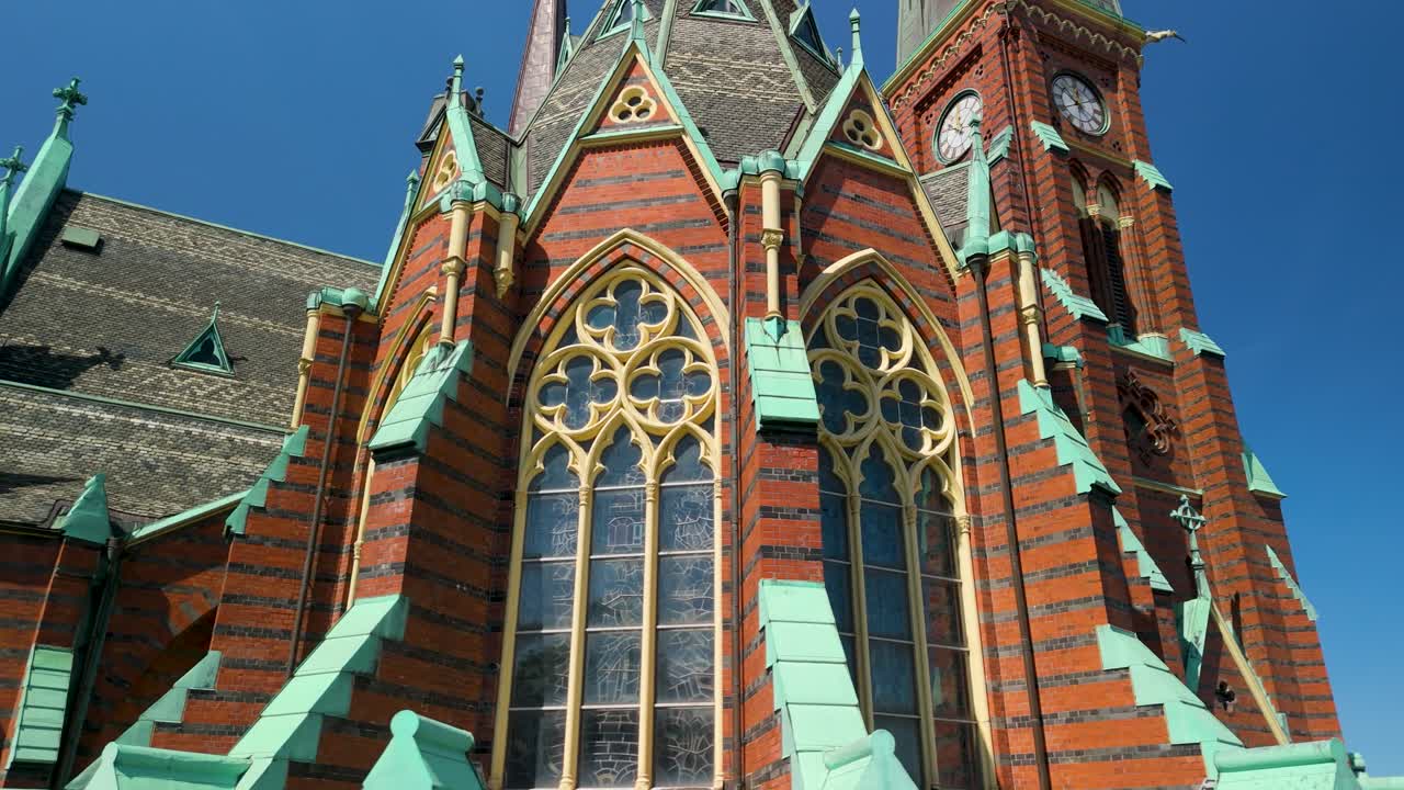 Drone afslører Oscar Fredriks Kyrkas udsmykkede nygotiske facade med intrikate spidsbuevinduer, guldfarvede buer, stribet murværk og irgrønne kobberdetaljer mod en blå himmel