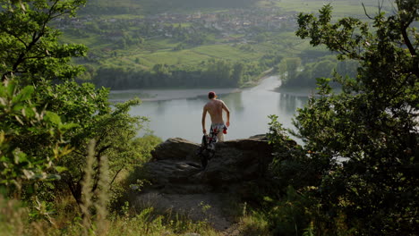 Hiker-taking-break-above-Danube-river,-wide-cinematic-shot,-austria,-durnstein,-europe,-hazy-mountain-valley-landscape,-independent-male-traveller,-sitting-down,-bare-chest,-wandering,-hike-trail