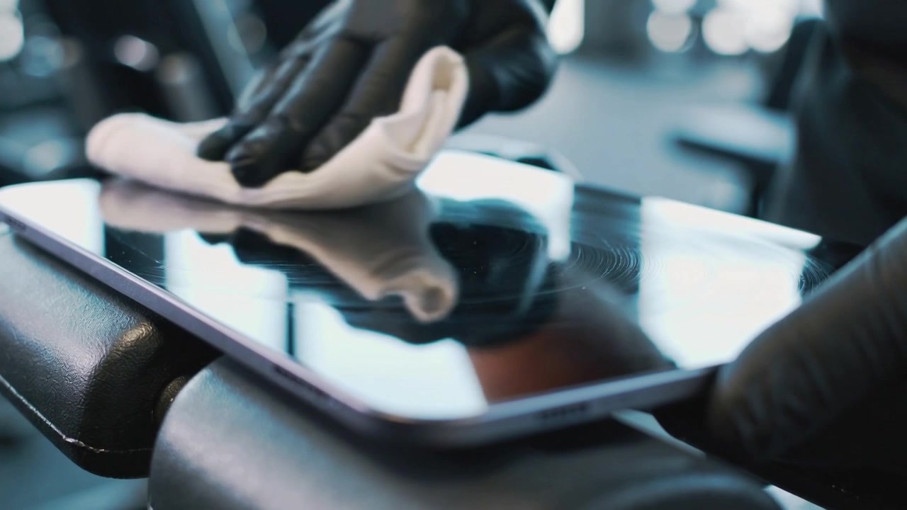 Nearing black-gloved hand pressing folded cloth wiping tablet on padded gym handle removing smudges