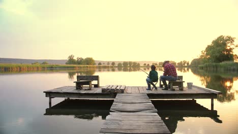 Rear-view-of-a-teen-boy-sitting-with-his-grandfather-on-the-lake-pier,-talking-and-fishing-together