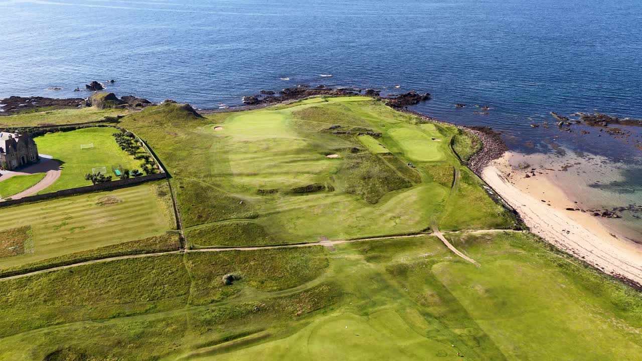 Drone glides above green golf course, sandy beach, and blue sea under bright daylight