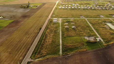 Private-homes-district-in-green-meadows-with-vehicles-driving-on-road,-aerial-golden-sunset-view