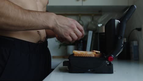 hombre camina hasta una prensa de parrilla de panini y coloca rebanadas de queso de mármol en dos pedazos de pan blanco, preparándose para hacer queso a la parrilla en su encimera blanca