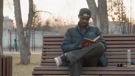 young man in casual winter clothing sits cross legged on wooden park bench reading hardcover book with deep focus, while background shows cyclist passing and bare trees in soft golden evening light