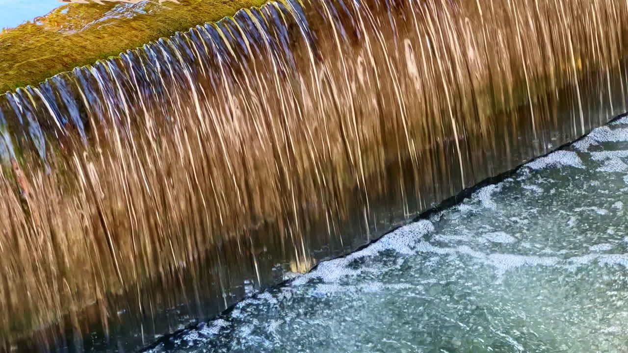 Anmutig stürzt Wasser über den Rand und erzeugt einen friedlichen Strom, der in einer idyllischen Flusslandschaft am Nachmittag im Sonnenlicht glitzert.