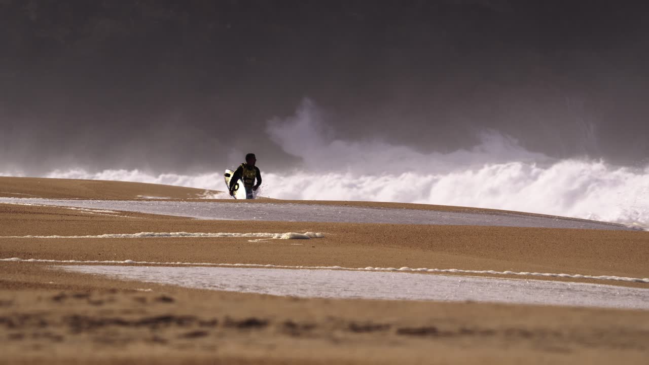 Mand går på en tom strand i våddragt, bærende et surfbræt.
