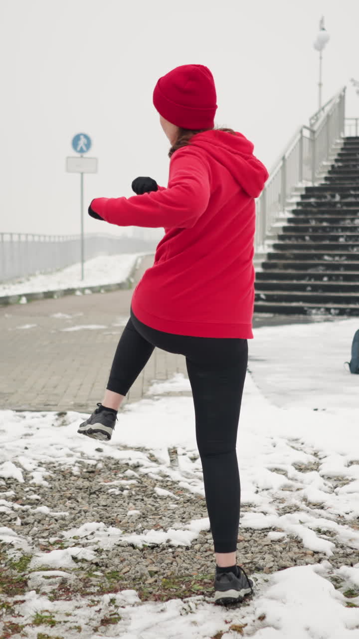back view of woman performing energetic winter workout outdoors on snowy ground near staircase and railing wearing red hoodie and black gloves demonstrating fitness moves