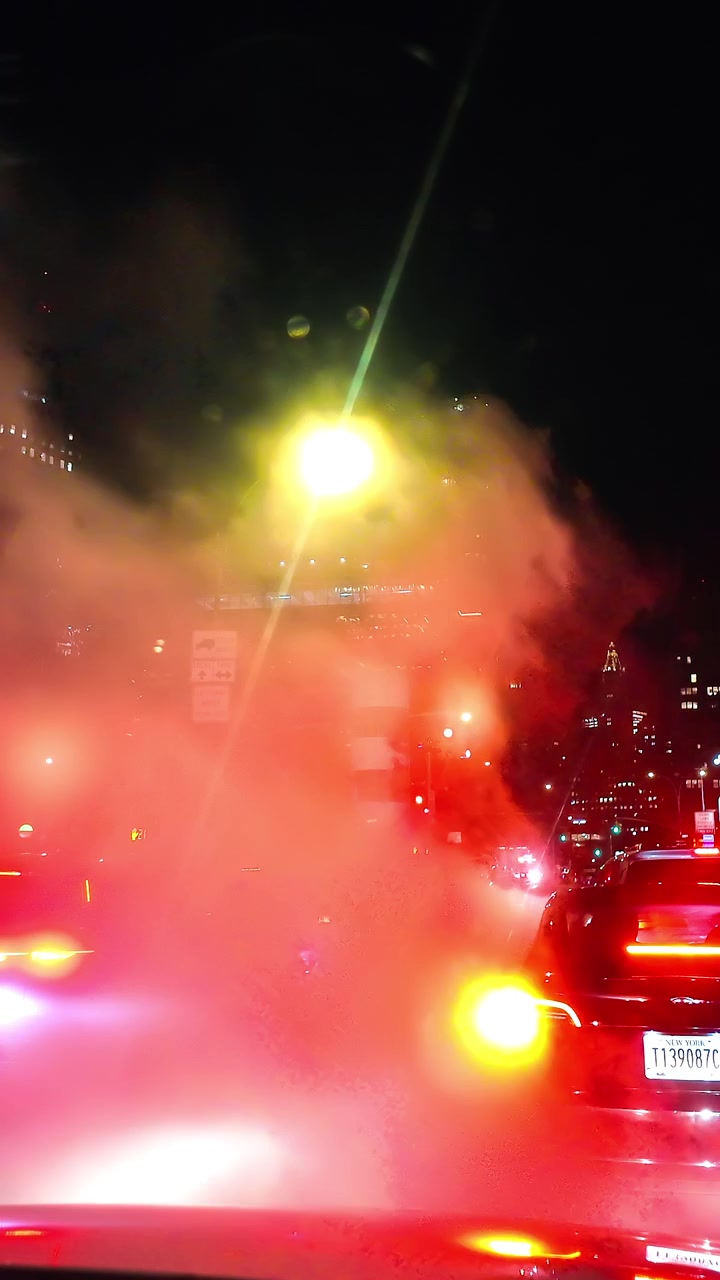 New York, USA, 25 December 2025: Steam rising from street vent on a night road, Manhattan, New York. Glowing taillights and streetlamp flare through vapor with city skyline in background.