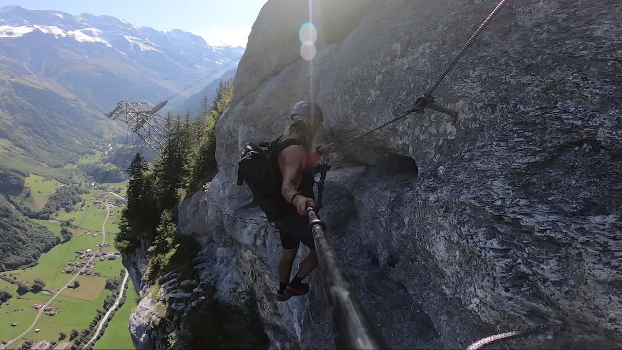 Athletic and fit man takes careful steps as he walks on the mountainside on Via Ferrata route in Switzerland. Clear sky with hot sunlight and epic landscape behind him in the valley.
