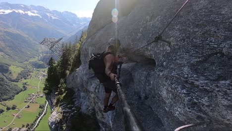 athletic and fit man takes careful steps as he walks on the mountainside on via ferrata route in switzerland. clear sky with hot sunlight and epic landscape behind him in the valley.