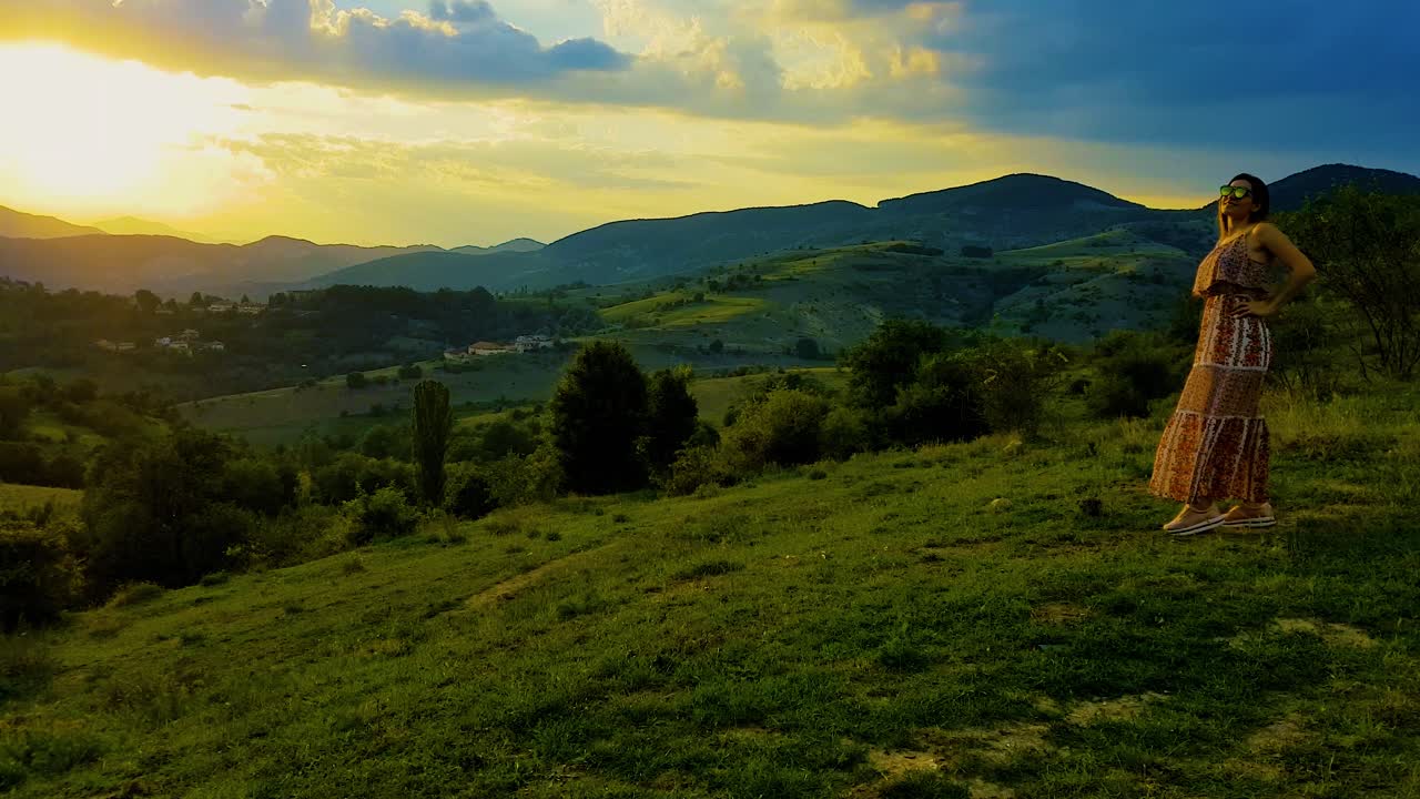 atardecer nublado sobre las montañas con una chica posando en una colina cubierta de hierba