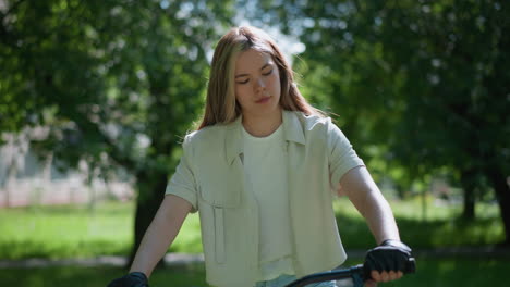 young woman wearing biker gloves sits thoughtfully on her bicycle in a green park, watching her hands while surrounded by nature, background features fluttering leaves and sun-dappled trees