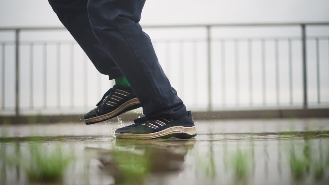 젖은 산책로, 신발, 다리가 클로즈업된 이미지예요. 생각에 잠긴 백인 남성 통근자가 난간 근처로 조심스럽게 다가가고, 질감이 있는 볼라드를 조심스럽게 밟고 피해 갑니다. 미끄러운 보도에서 균형을 잡으며 잠시 멈추고, 반사되는 물웅덩이에 모습이 비치네요.
