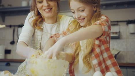 young beautiful mother teaching her pretty daughter to cook and knead a daugh for cookies on the table in the kitchen. inside