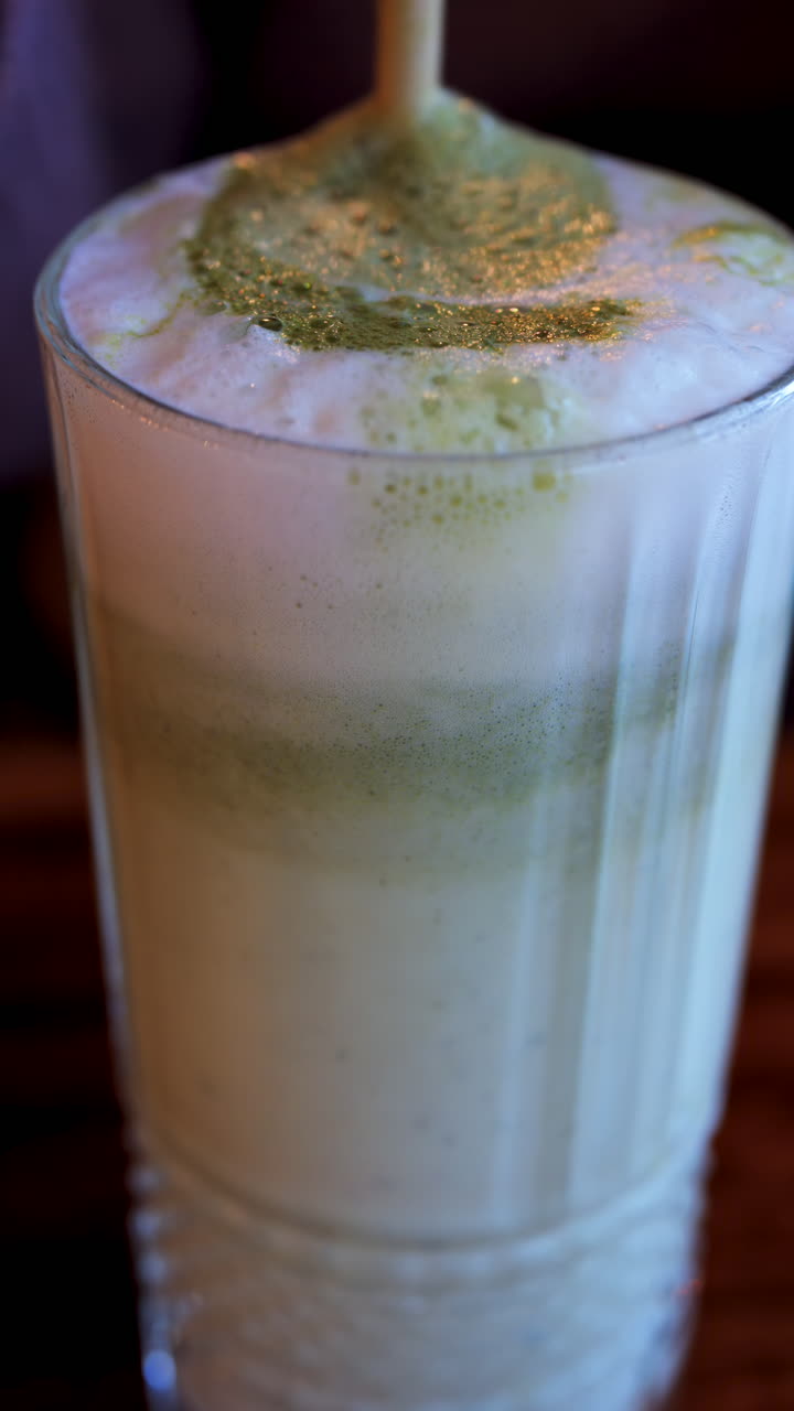 Close up of a straw mixing in a matcha latte on a table at a cafe. Vertical