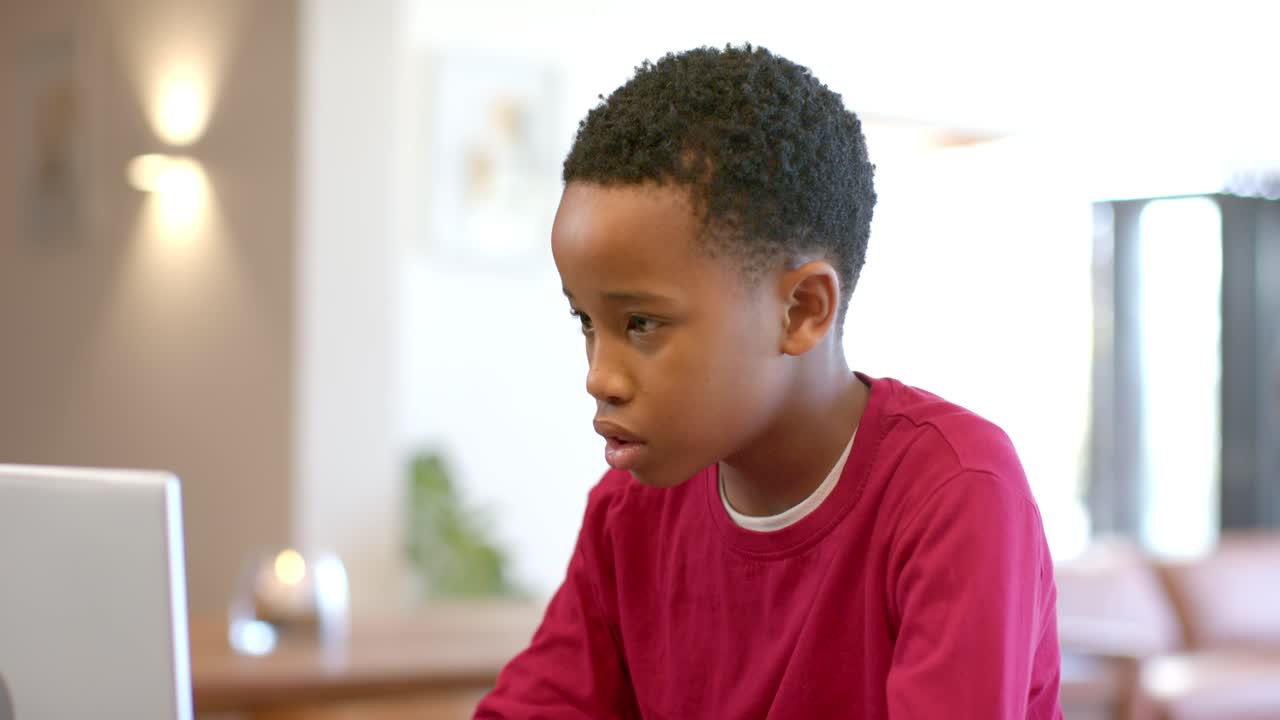 Ragazzo afroamericano che preme i tasti, fa i compiti al tavolo con il padre che usa lo smartphone sullo sfondo