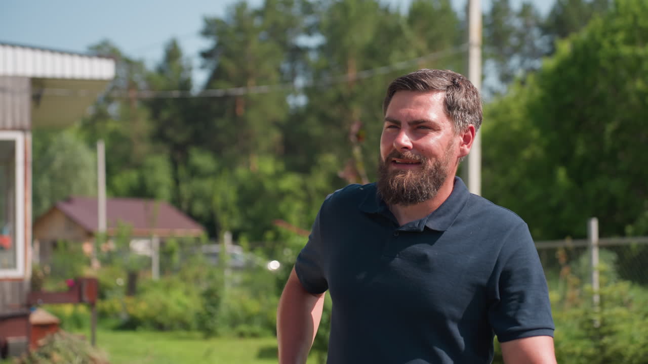Young farmer walking toward farm holding plier, pauses with subtle smile while looking around, sunlight on grass and plant beds, brick house and trees in background