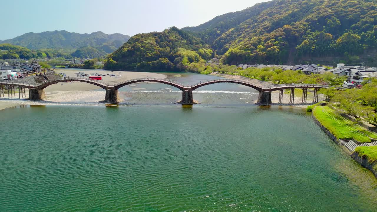 An aerial shot trucks right, following the iconic Kintai Bridge in Iwakuni, Japan. The historic wooden arches span the Nishiki River, with lush green mountains and a town in the background