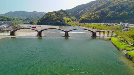 an aerial shot trucks right, following the iconic kintai bridge in iwakuni, japan. the historic wooden arches span the nishiki river, with lush green mountains and a town in the background
