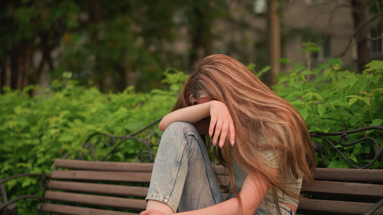 Young woman in jeans and white sneakers sits on wooden bench in park, resting head on arm in reflective, somber posture with polished nails and long hair flowing over her shoulder