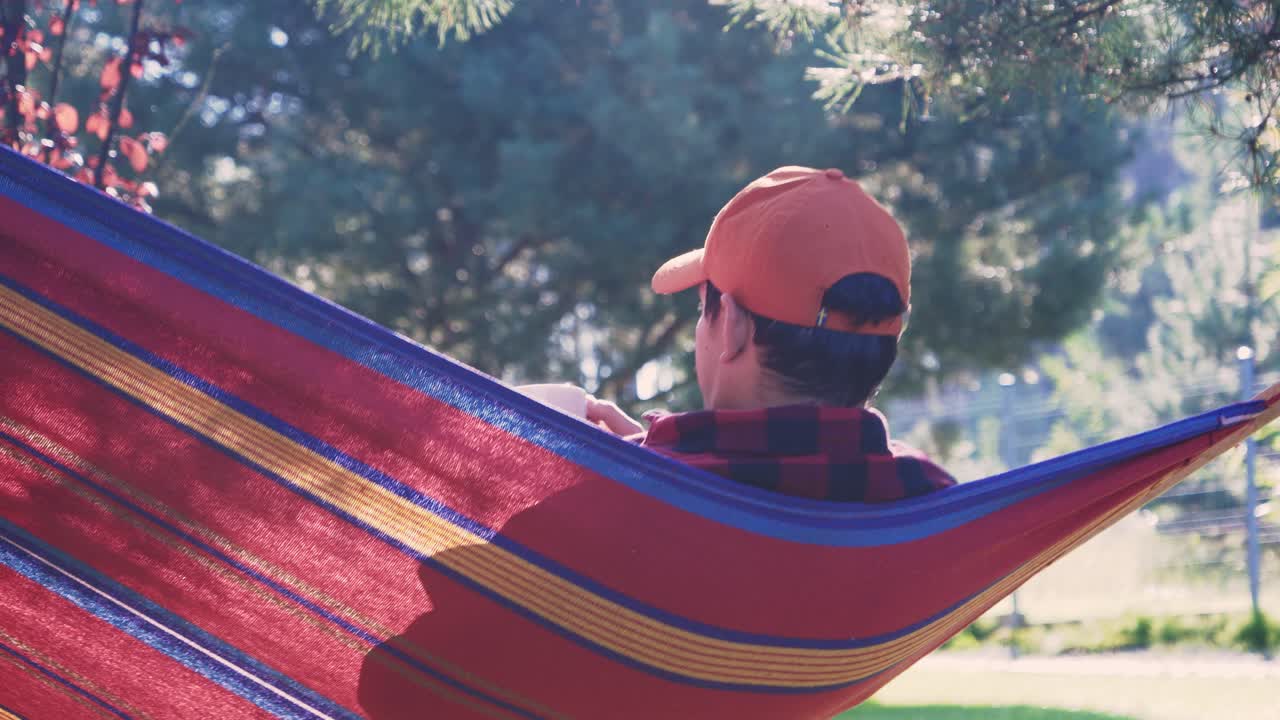 hombre relajándose en una hamaca mirando lejos mientras bebe una bebida de café caliente