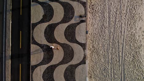 Top-down-aerial-split-view-of-Copacabana-beach-with-patterns-in-the-sand-on-the-right-and-the-pavement-boulevard-on-the-left