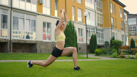 Una-Mujer-Joven-En-El-Parque-Con-Auriculares-Hace-Yoga.-Haciendo-Yoga-En-El-Césped-Con-El-Telón-De-Fondo-De-Casas-En-El-Parque-De-La-Ciudad-En-Cámara-Lenta