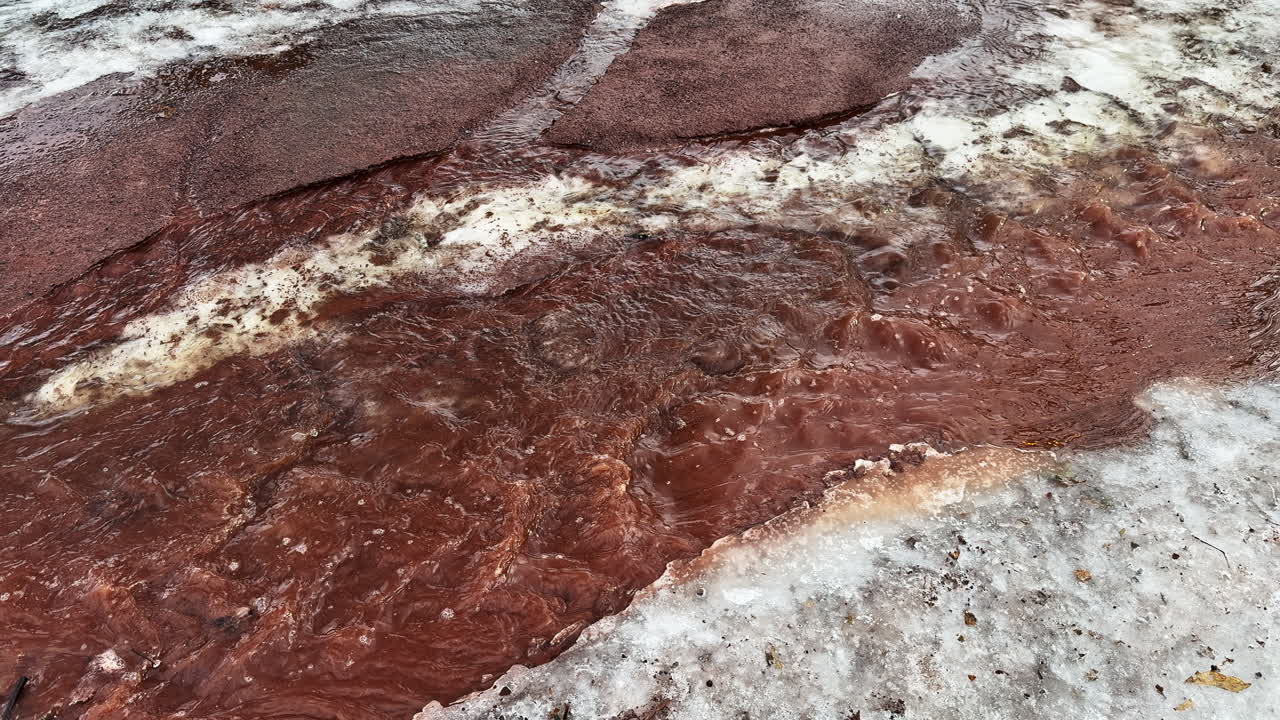 Abbondante acqua di disgelo turbina tra neve persistente e terra rossa che si sta scongelando, creando un paesaggio di fine inverno ricco di texture dove la primavera inizia a farsi strada.