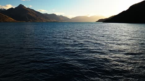 Plataforma-Rodante-Aérea-Sobre-El-Agua-Del-Lago-Wakatipu-Ondulaciones-En-La-Superficie-Al-Atardecer-Con-Destellos-De-Montaña