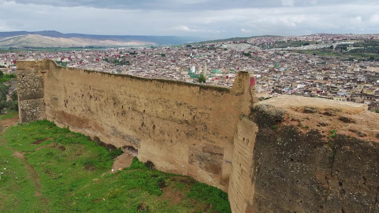 Vista aerea lungo la storica cinta muraria fortificata di Fez, Marocco, con architettura difensiva invecchiata in primo piano e il denso paesaggio urbano che si estende sulle colline oltre.