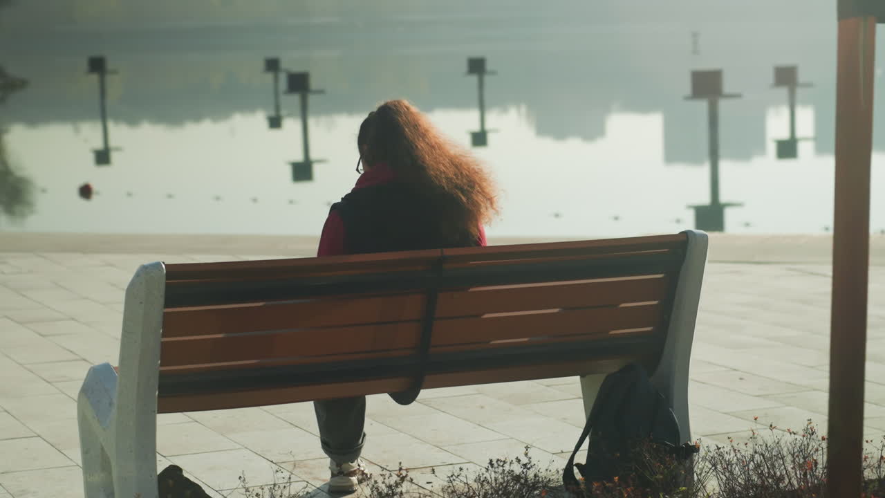 Solitary person on bench by waterfront, curly hair profile in late morning sunlight creating soft reflection on calm harbor pier posts and distant signage frame scene, bin and bag at feet, cinematic