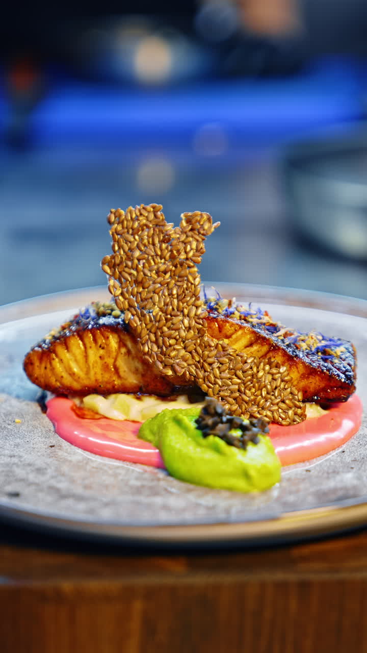 A beautiful dish of fried salmon with two sauces. Cook's hand puts the chips made of sesame seeds on the plate. Close up. Blurred backdrop. Vertical video.