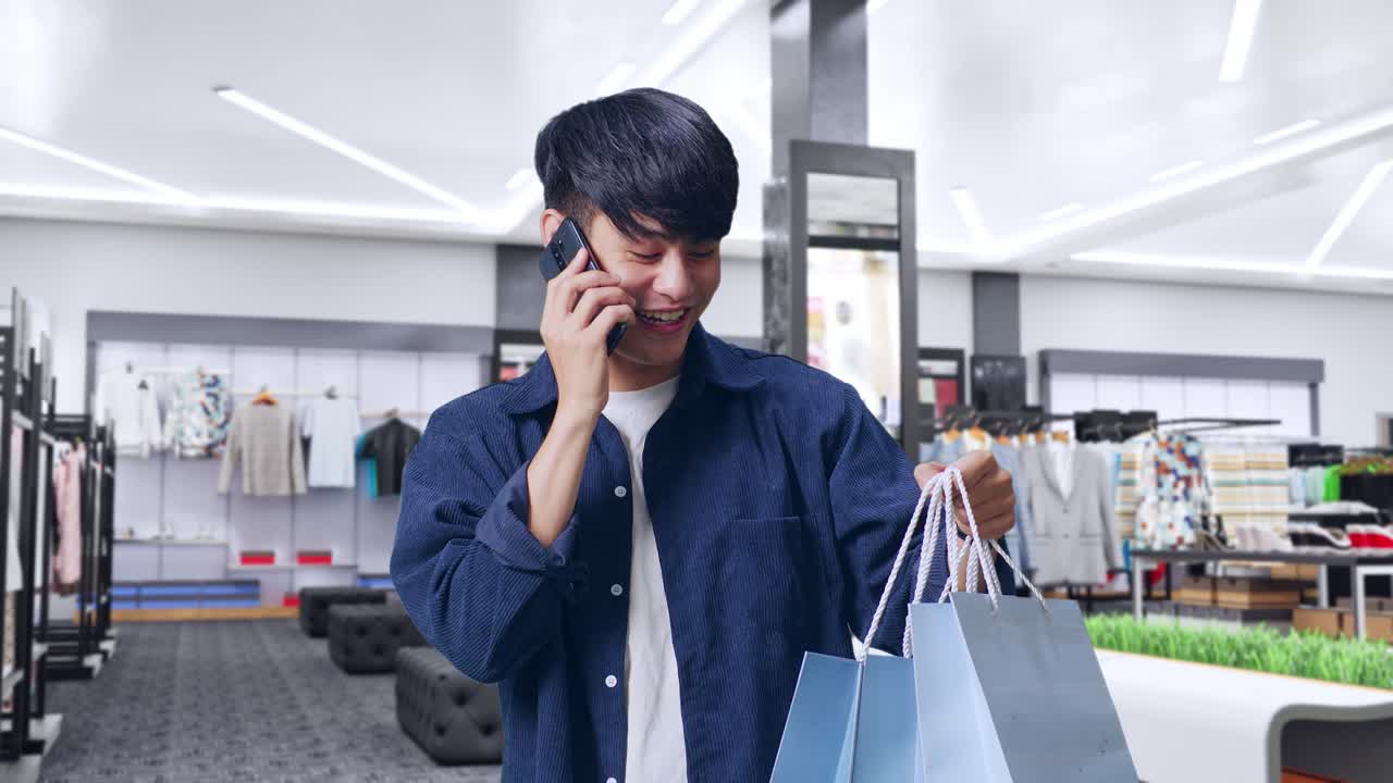 옷 가게 에 서 있는 동안 전화 에 응답 하고 휴대 전화 에 대화 하는 쇼핑 가방 을 가진 아시아인 쇼핑 남자