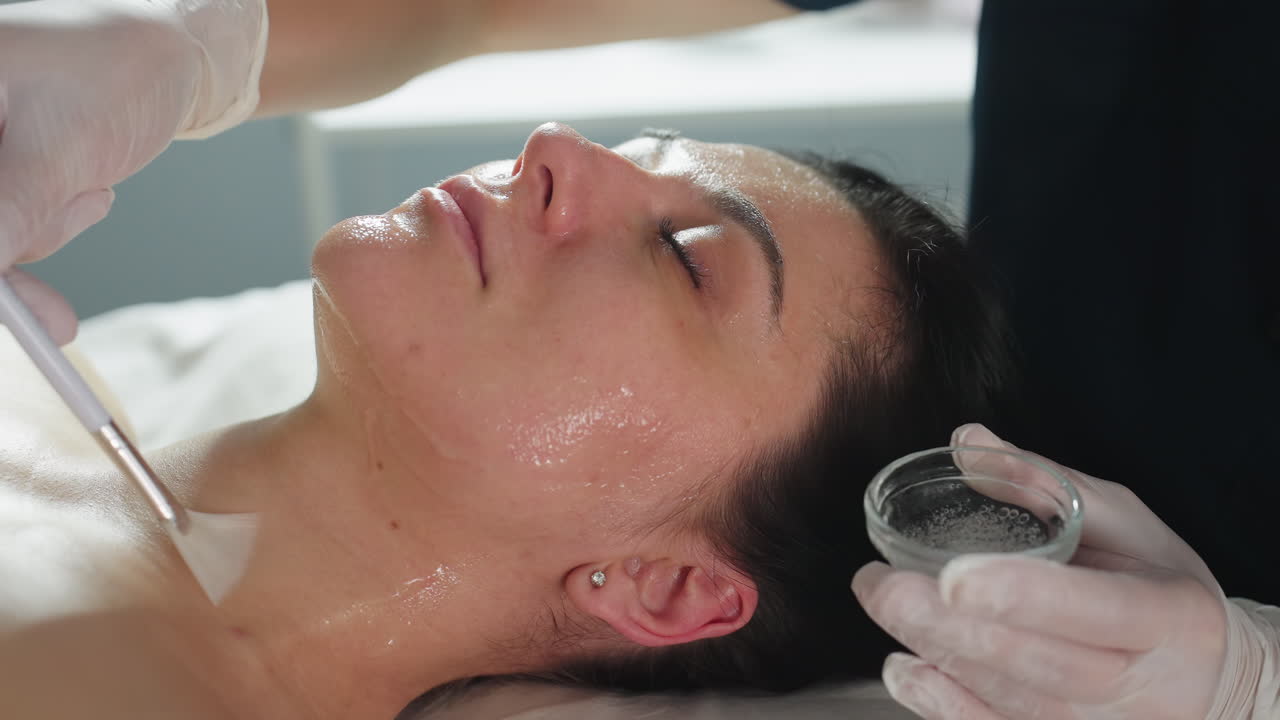 Female groomer in gloves applies clear facial massage gel with brush from chin up to neck on relaxed client lying on spa bed under soft clinical light for nourishing skincare treatment session
