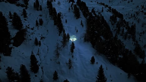 Vista-Aérea-De-Un-Excursionista-Solitario,-Iluminado-Por-Un-Faro,-Caminando-Por-Un-Paisaje-Montañoso-Nevado-Por-La-Noche