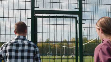 young people viewed from behind entering a volleyball court as the coach opens the gate, framed by a football field and lush trees in the background