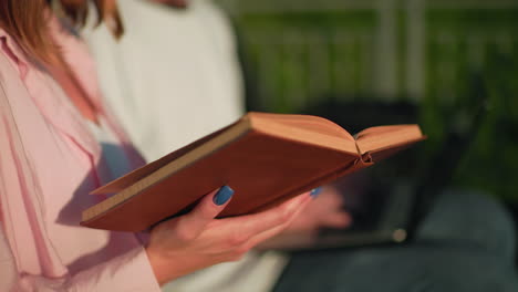 primer plano de una mujer en rosa leyendo un libro con uñas pintadas de azul mientras su amiga en el fondo escribe en una computadora portátil, ligeramente borrosa, la escena incluye una colina verde, un poste y un entorno al aire libre relajado