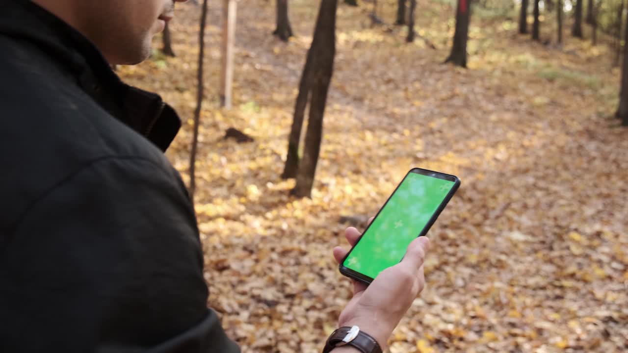 joven sostiene el teléfono móvil con pantalla verde chromakey seguimiento mate en el parque del bosque de otoño