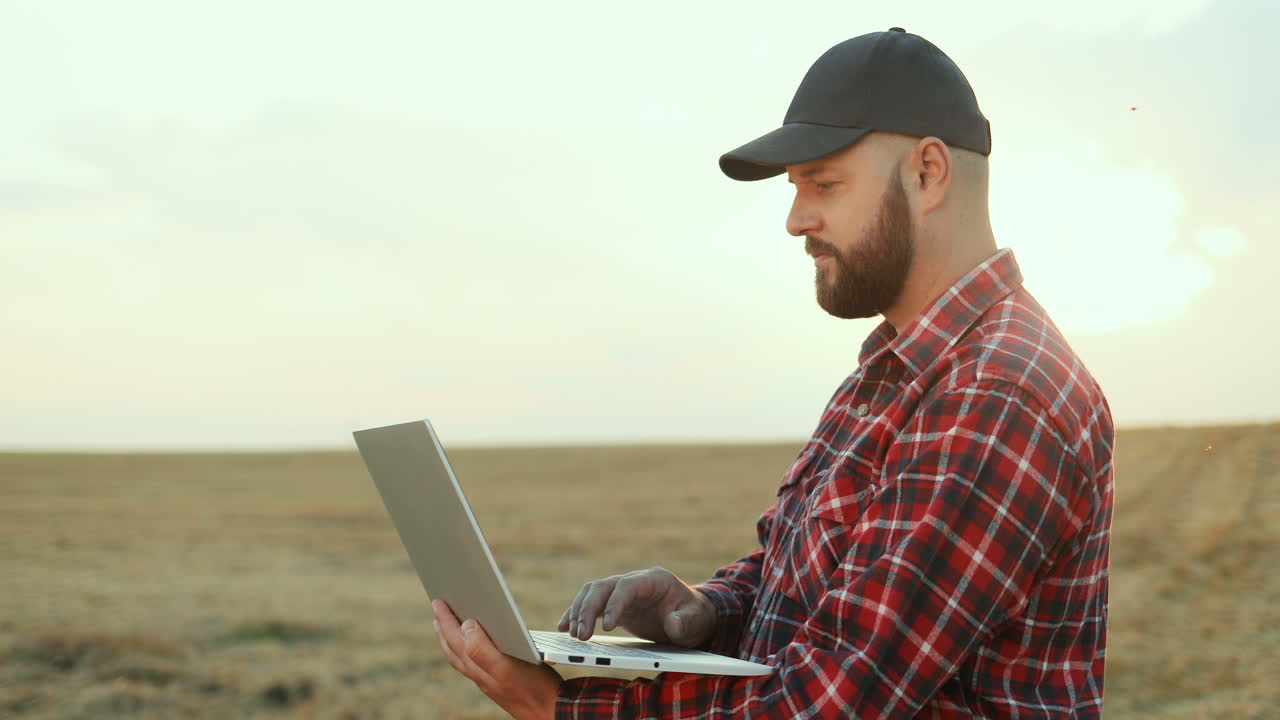 Close up of farmer wearing a shirt and black caseball cap looking at the sides and thiking while working on laptop computer