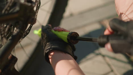 primer plano de manos con guantes bombeando aire intensamente en el neumático de la bicicleta usando una boquilla verde en un día soleado, el fondo suavemente borroso, mostrando detalles sutiles de los radios y el pavimento