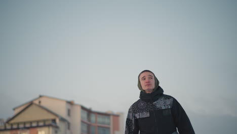 upward view of young boy skating outdoors in patterned winter jacket under bright open sky with faint outline of distant building and soft evening light creating calm winter scene