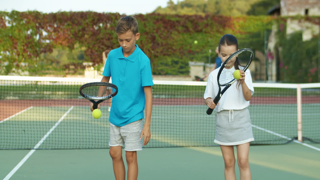 Sister And Brother Playing At Tennis Court And Hitting Balls Against The Ground With Racket