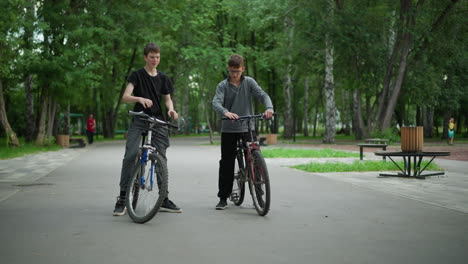 two brothers on bicycles share a friendly moment in a park, each greet themselves with a knuckle bump with a smile, the background showcases lush green trees