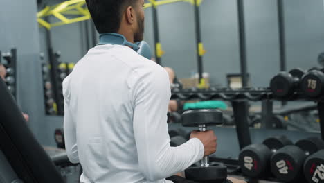 side view of gym rat in white shirt sitting and placing dumbbell on leg inside well equipped gym with visible dumbbells and racks, another gym-goer passes by in background